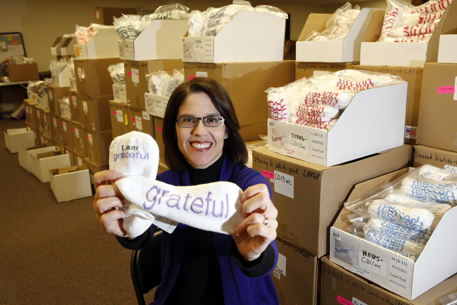 A picture of an SBDC client who sells special socks. The client is in the foreground holding a pair of socks, and in the background are numerous boxes with socks packaged inside.